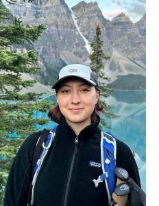 Portrait of IDEA Grant recipient Rebecca Denhart - student is smiling, has a cap on her head, and is wearing a black jacket, there are mountains and a lake in the background.