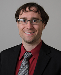 Alexander Dupuy, a faculty member with brown hair and glasses, is looking at the camera.